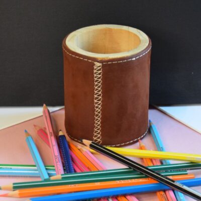 Cette photo représente un étui à crayon en bois recouvert de cuir marron. Des crayons sont posés à côté